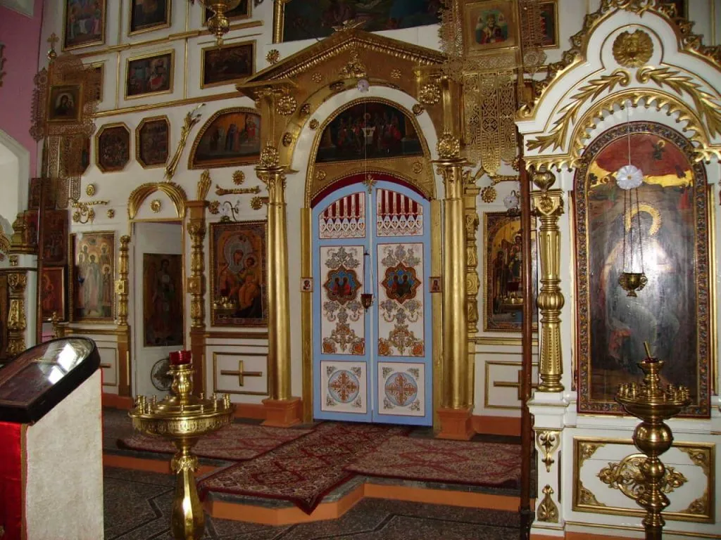 Иконный интерьер православной церкви с золотыми деталями и богато украшенными дверями. Внутри храма видны иконы, светильники и свечи, создающие атмосферу духовности и спокойствия.