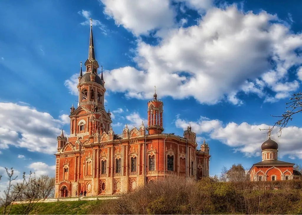 Красивая церковь в Подмосковье на фоне голубого неба с облаками. Архитектура в стиле барокко, ярко-красный кирпич и золотые детали создают уникальный облик здания. Идеально для фото и туристических прогулок.