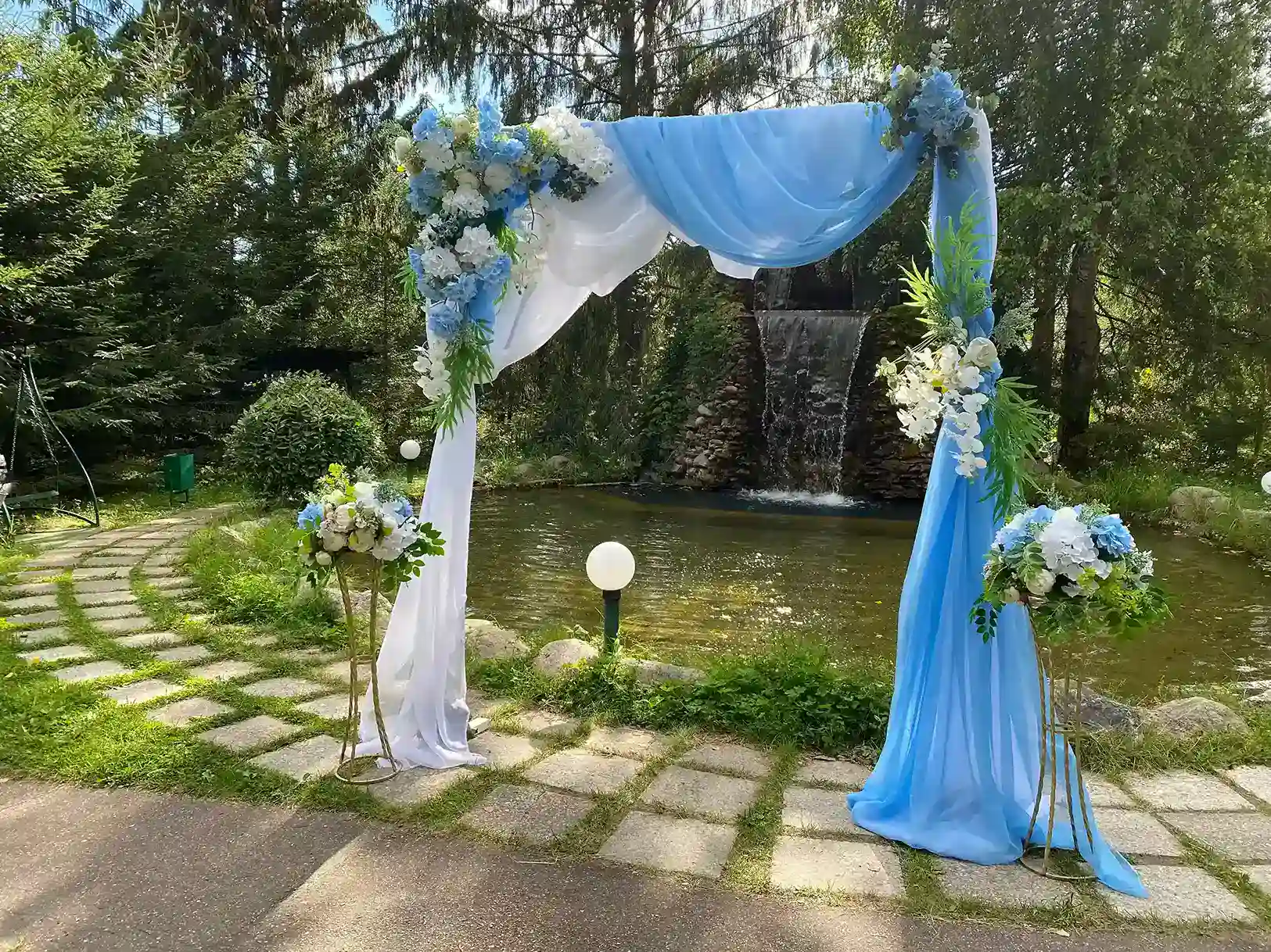 Красивая свадебная арка, украшенная голубыми и белыми цветами, установленная на берегу пруда с водопадом, окруженная зеленью и деревьями в живописном природном месте.