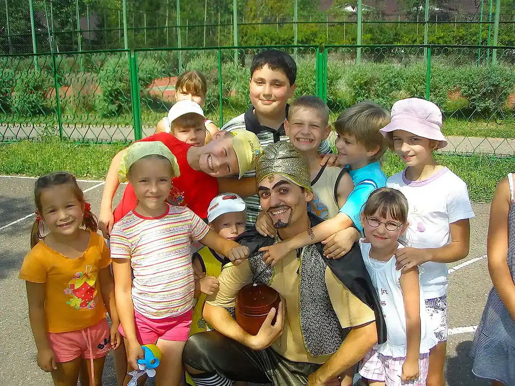 Веселая компания детей и двух взрослых на улице, на заднем плане забор и зелень, яркие летние наряды, улыбки и дружелюбная атмосфера.