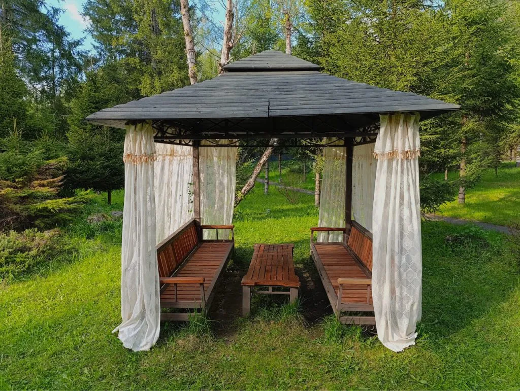 Уютная беседка с деревянными скамейками и шторами в зеленом парке, идеально подходит для отдыха на природе в Подмосковье. Отличное место для отдыха и общения на свежем воздухе.