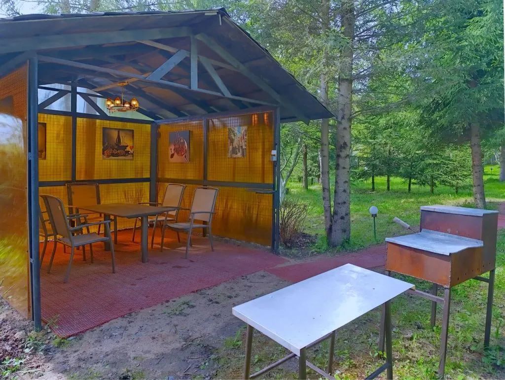 Уличная беседка с столом и стульями на природе среди деревьев, уютное место для отдыха и барбекю. Освещена декоративным светильником, окружена зеленью и создаёт атмосферу спокойствия и уединения.