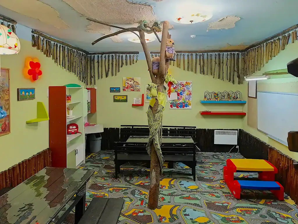 Яркий детский игровой зал с деревьями и светлыми стенами, разнообразными игрушками, картинами и декоративными элементами, создает веселую и уютную атмосферу для малышей.