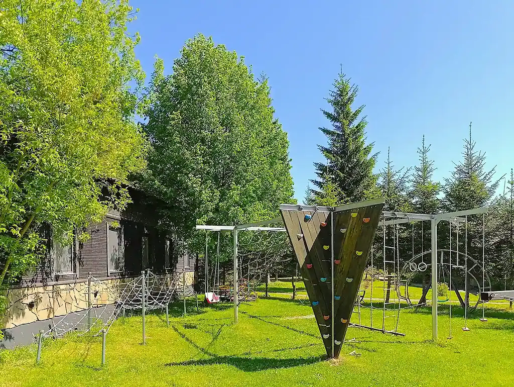 Красивый зеленый сад с игровой зоной, включающей скалодром и веревочные конструкции, окруженный деревьями и уютным домом, отлично подходит для отдыха и развлечений на природе.