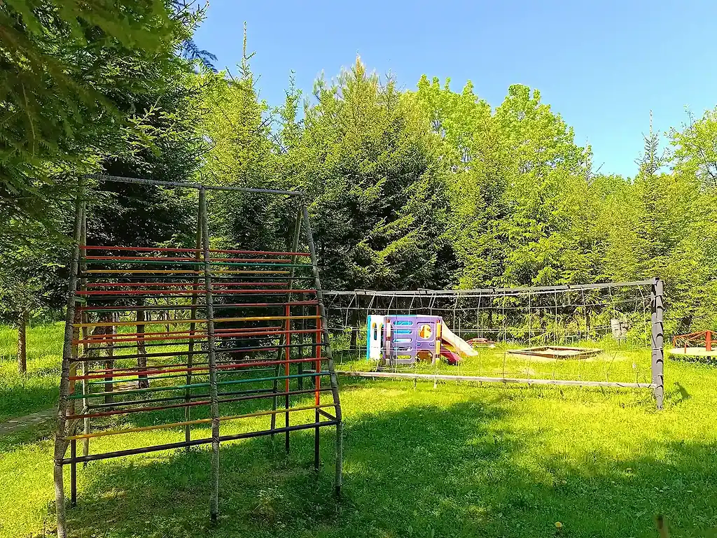 Детская игровая площадка на зеленой территории с деревьями, включает лесенку, сетку и пластиковую горку, идеально подходит для отдыха в природе в ВЕ Art Resort.