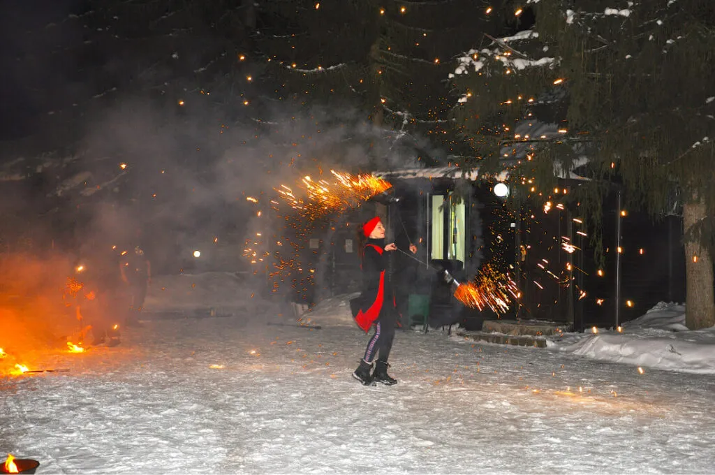 О девушка катается на коньках и зажигает бенгальские огни в зимнем лесу у деревянного дома, создавая яркое зрелище на снегу.