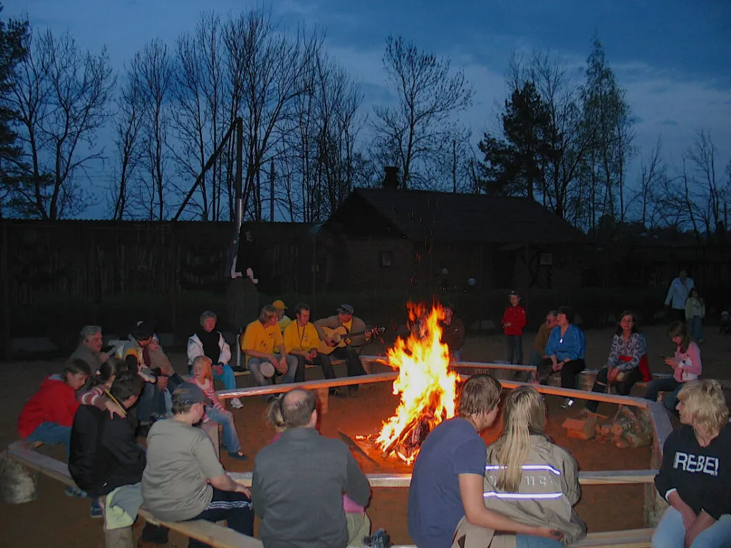 яркое костровище с людьми на вечерней вечеринке в лесу, группа гостей у огня, играет музыка, деревья и дом на фоне, отдых в Подмосковье.