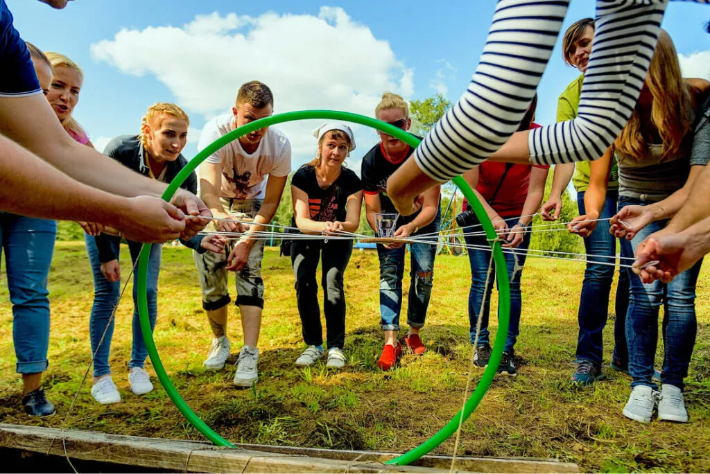 Экипажное мероприятие на свежем воздухе, участники участвуют в командной игре с нитями на зеленом поле под ярким голубым небом.