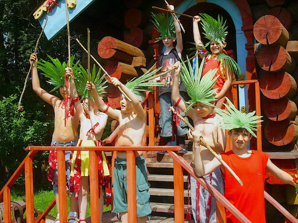 Летние дети в традиционных пляжных костюмах с листьями и палками, играющие на деревянной площадке в природном окружении.