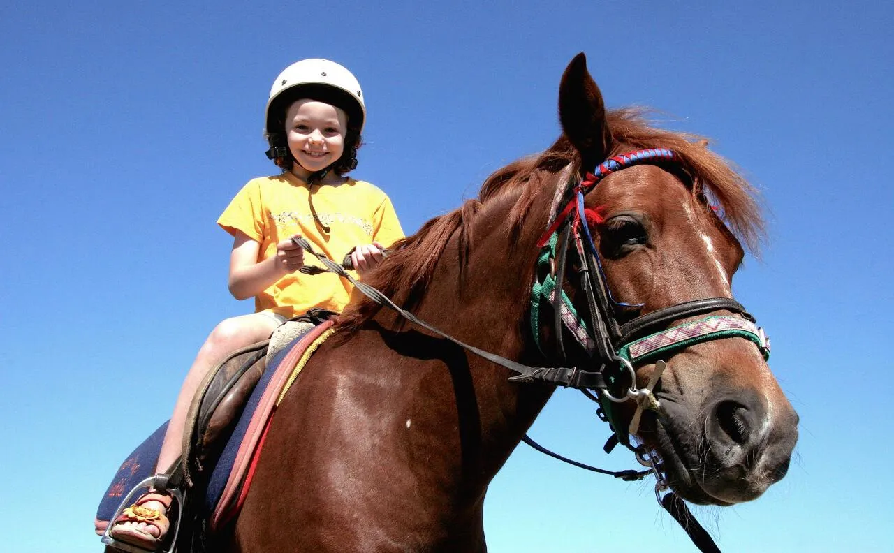 Улыбающаяся девочка в шлеме едет верхом на лошади на ярком солнечном небе.