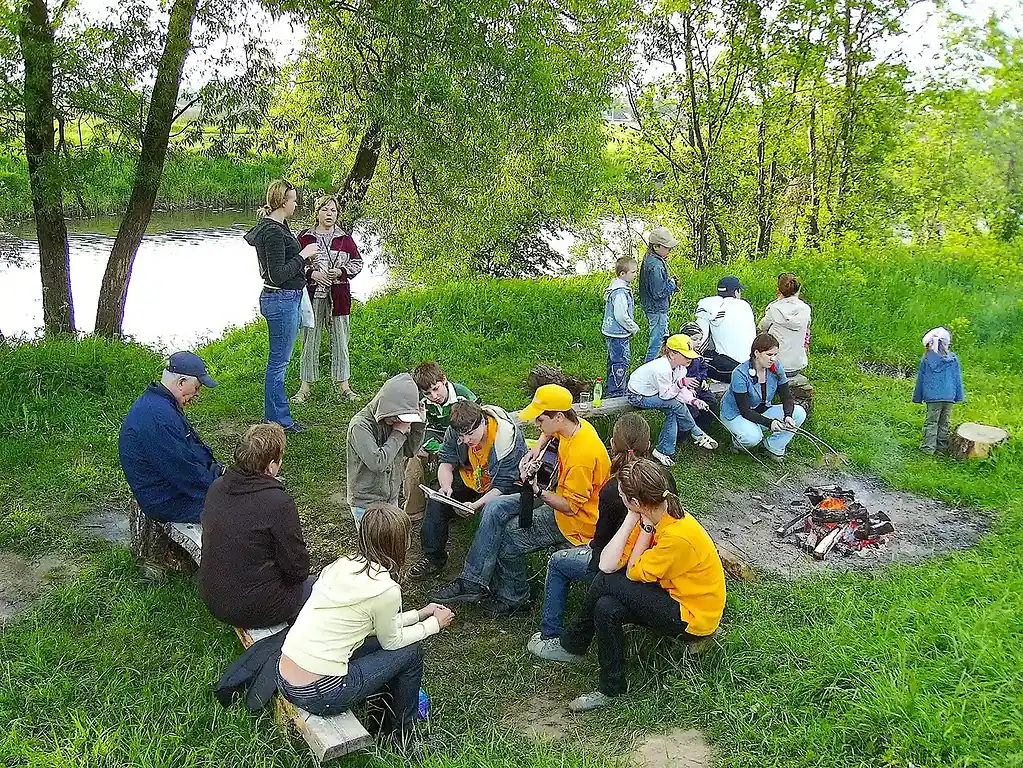 Летний отдых на природе у водоема с группой людей, пикник и посиделки у костра в окружении деревьев в Подмосковье.