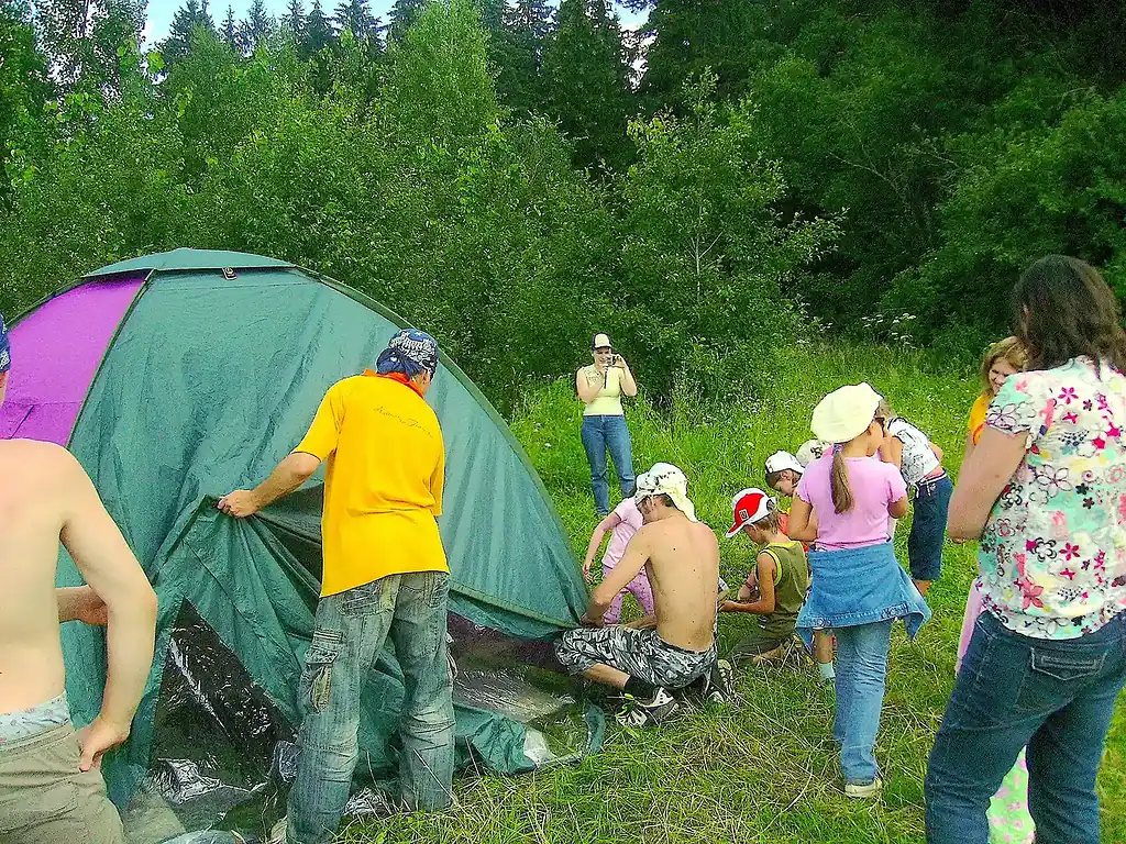 Ребята собирают палатку на природе, в окружении зелени и деревьев, яркое теплое лето, веселый отдых на свежем воздухе.
