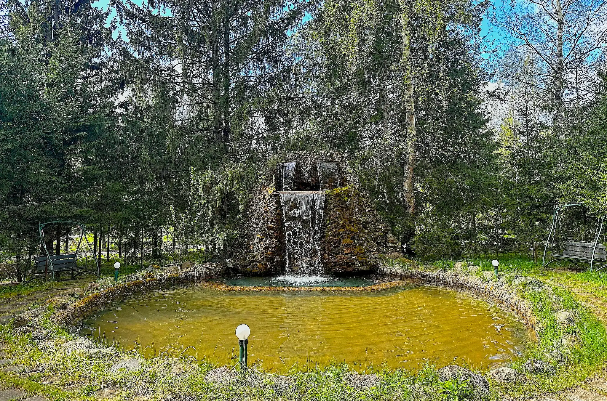 Уголок природы с водопадом и прудом, окруженный соснами и березами, подходит для отдыха на свежем воздухе, спокойствия и уединения в лесу.